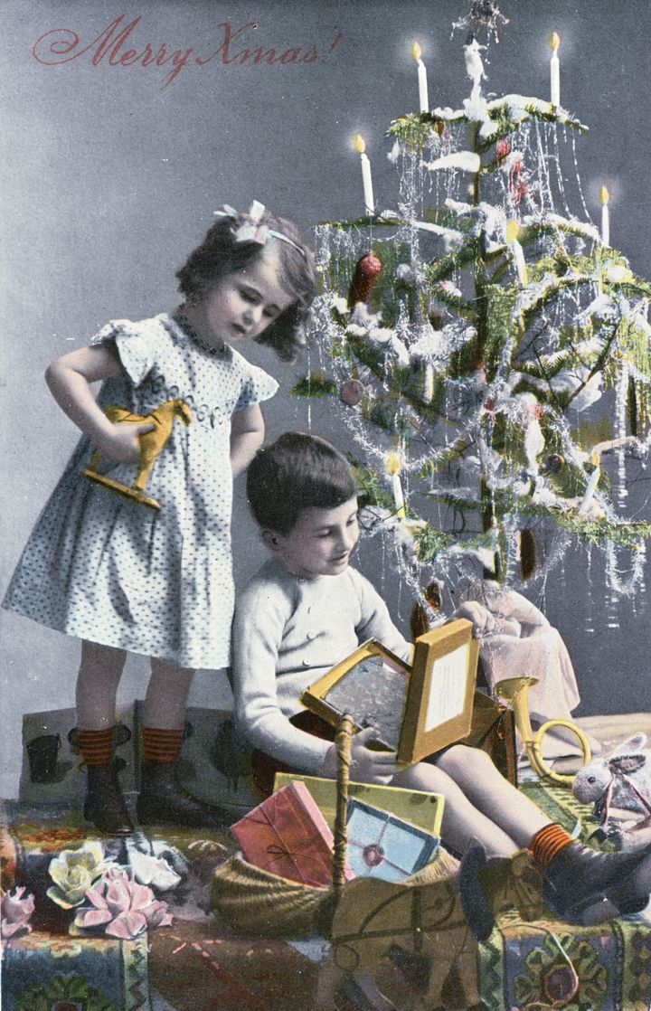 Επιχρωματισμένη φωτογραφία (τέλη 1800) με δύο παιδιά, ένα κορίτσι και ένα αγόρι που ανοίγουν τα δώρα τους πλάι σε χριστουγεννιάτικο δέντρο, το οποίο βρίσκεται πάνω σε τραπέζι και είναι φωτισμένο με κεριά.