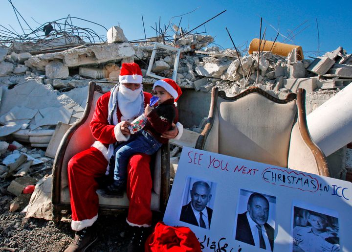 Παλαιστίνιος ντυμένος Άγιος Βασίλης κάθεται σε μια πολυθρόνα στα ερείπια σπιτιού ομοεθνή του που κατεδάφισαν οι Ισραηλινοί. Στα πόδια του έχει ένα αγοράκι. Χωριό αλ Καντέρ, δυτικά τη Βηθελεέμ