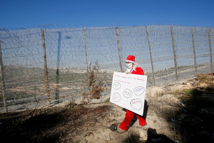 Παλαιστίνιος διαδηλωτής ντυμένος Άγιος Βασίλης κρατά ένα πλακάτ και κινείται πλάι στον σιδερόφραχτο φράχτη κοντά στην Βηθλεέμ 