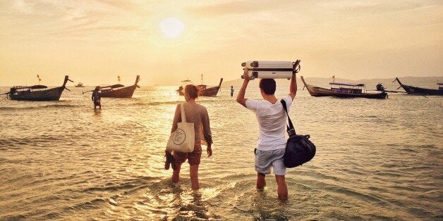 Young couple at Ao Nang beach in Krabi province walking into the ocean to long tail boats during low tide and holding their bags over their heads.