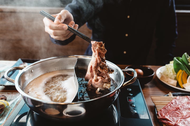 Man holding medium rare slice Wagyu A5 beef out from hot pot shabu shoyu soup base by chopsticks with steam.