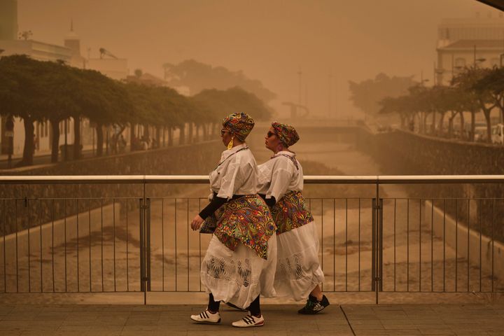 Two people in carnival dress walk across a bridge in a cloud of red dust in Santa Cruz de Tenerife.