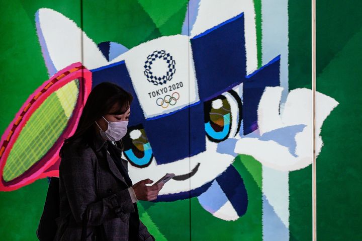 A woman wearing a face mask walks in the tunnel of a metro station with a poster of Miraitowa, official mascot of the 2020 Summer Olympics in Tokyo on March 11, 2020. 