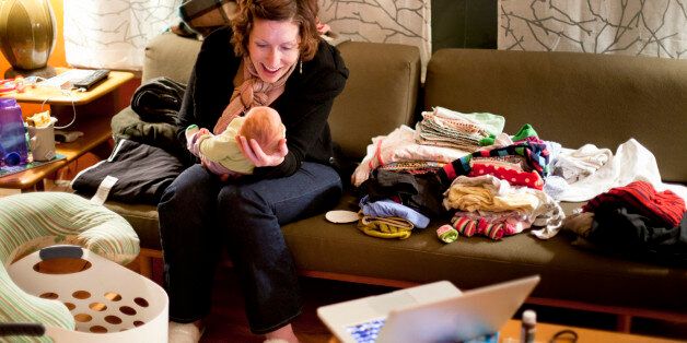 A smiling mother with red hair plays with her baby on a couch amid piles of messy laundry at night in a home.