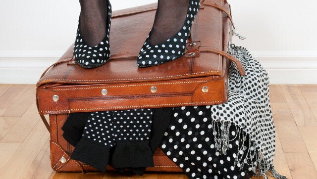 Woman in high heel shoes standing on leather suitcase overfilled with fashion clothing.