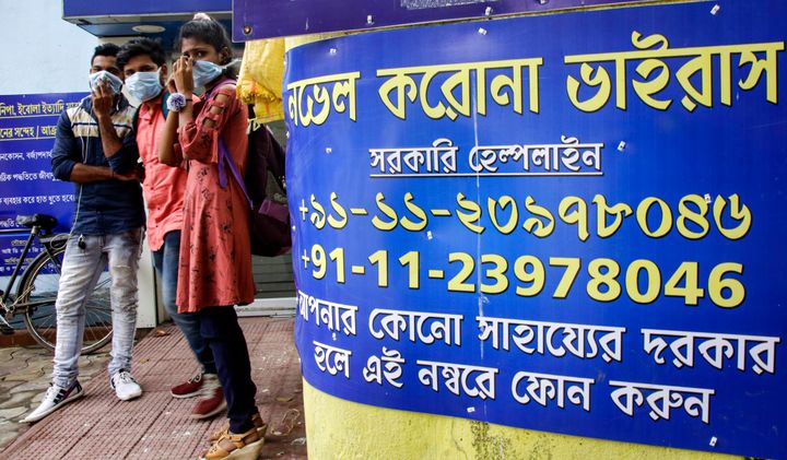 People wearing masks stand next to COVID-19 helpline number displayed on a wall at government run hospital in Kolkata, Friday, March 6, 2020.