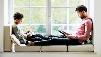 A father and son are relaxing by a large window at home, reading and playing games together.