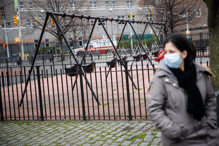 A woman wearing personal protective equipment sits alone alongside empty swings to maintain social distancing at a playground