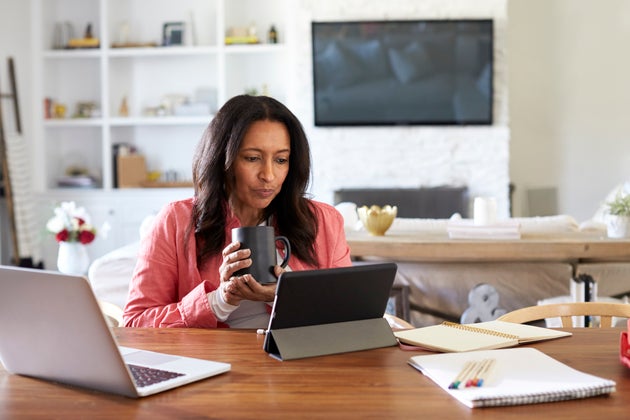 Middle aged woman sitting at a table reading using a tablet computer, holding a cup, front view