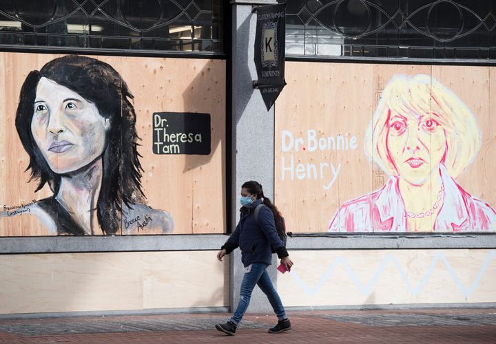 A woman wearing a protective face mask walks past portraits of Dr. Theresa Tam and Dr. Bonnie Henry on a boarded up business in downtown Vancouver, B.C. on April 1, 2020. 