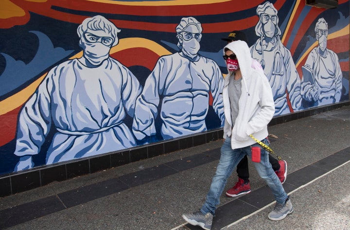 A man wearing a protective face mask walks past a mural of heath care workers painted on a boarded up restaurant in downtown Vancouver on April 27, 2020.