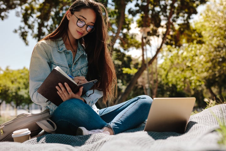 Close up of a college student sitting in a park and writing in her notebook. College student making notes looking at her laptop computer.