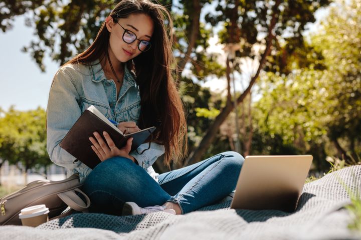 Close up of a college student sitting in a park and writing in her notebook. College student making notes looking at her laptop computer.