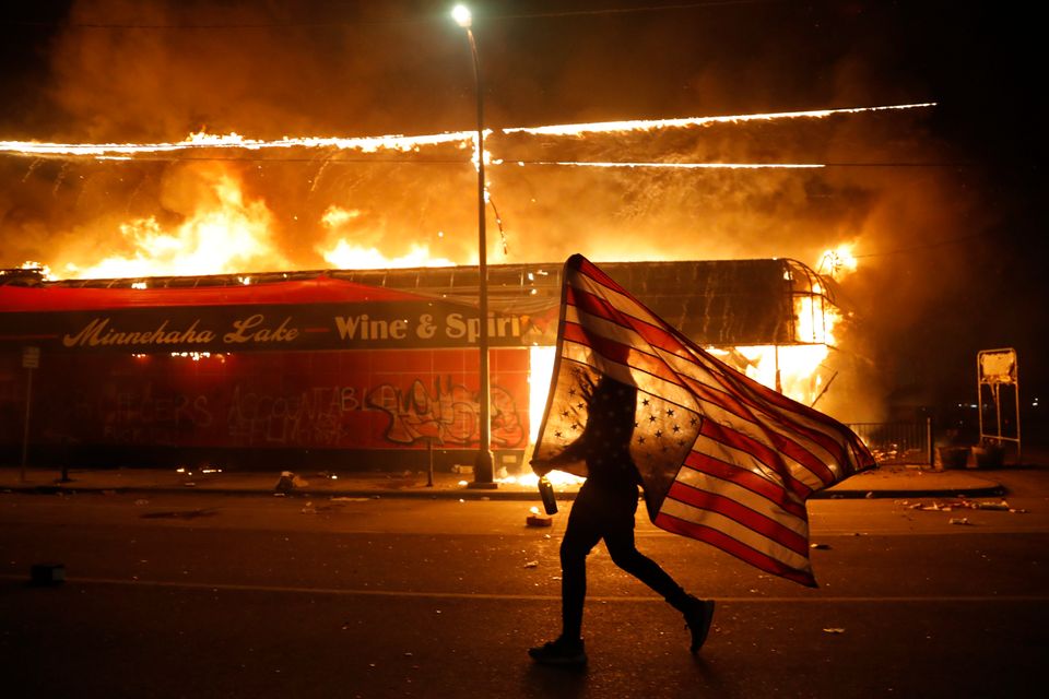 Διαδηλωτής κρατά την αμερικανική σημεία έξω από ένα φλεγόμενο μαγαζί, στη Μινεάπολη (AP Photo/Julio Cortez)