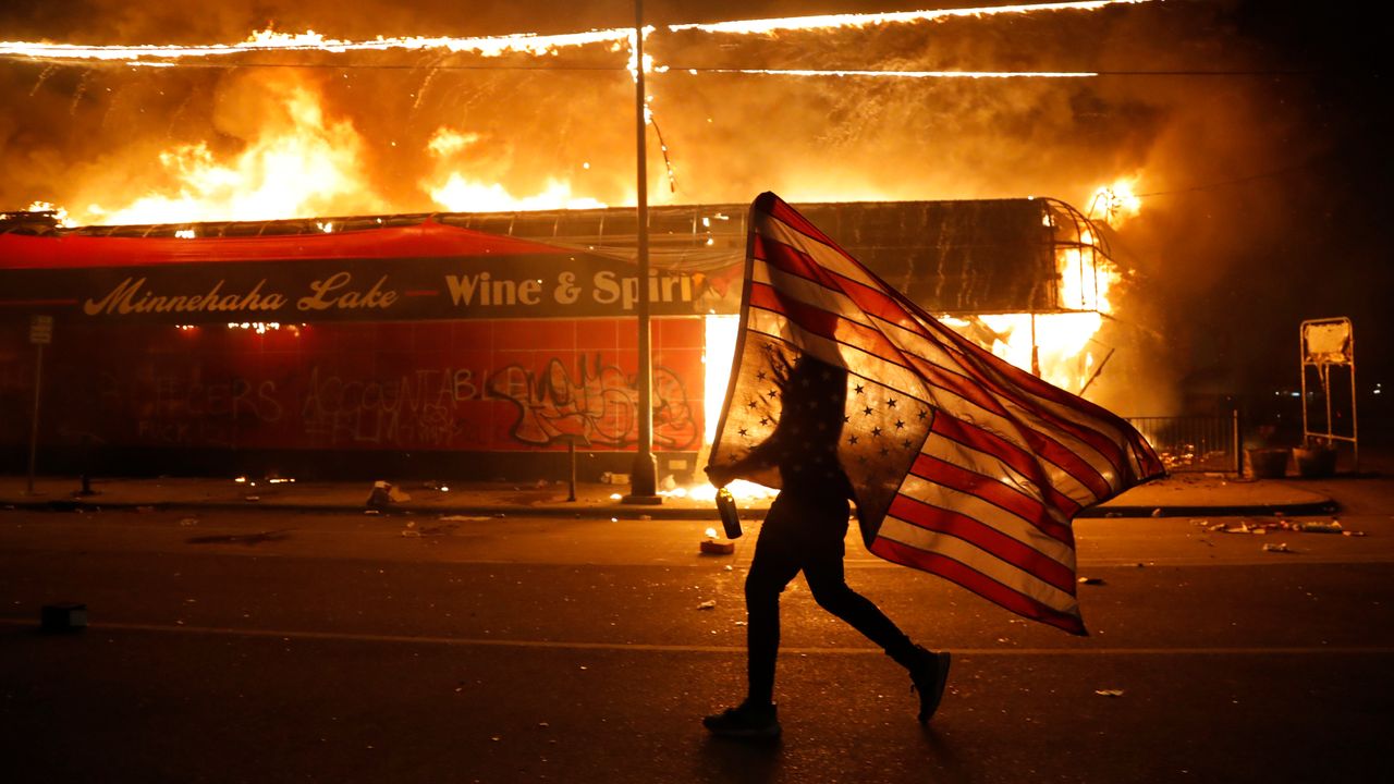 A protester carries the carries a U.S. flag upside, a sign of distress, next to a burning building on Thursday in Minneapolis. Protests over the death of George Floyd, a black man who died in police custody on May 25, broke out across the country over the weekend.