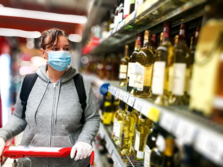 Selective focus color image depicting a caucasian woman in her 30s wearing a protective surgical face mask and plastic surgical gloves during the coronavirus (Covid-19) pandemic, in a bid to stop the spread of the virus. The woman is pushing her shopping cart inside a supermarket while shopping for wine and alcohol in the alcoholic drinks section of the store. Room for copy space.