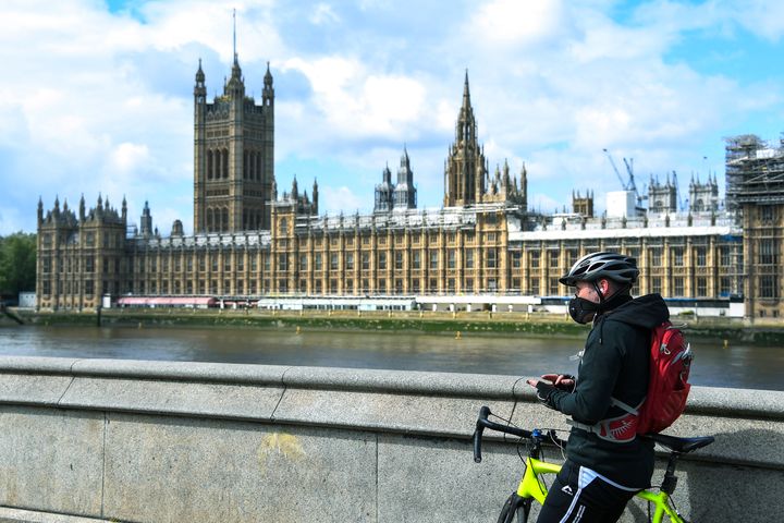 In this file photo dated Friday, May 1, 2020, a man stands with his bike, wearing a protective mask to protect against coronavirus, on the south bank of the River Thames, against the backdrop of the Houses of Parliament, in London.