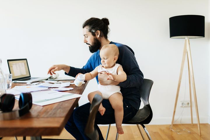 A single father working at his desk at home while holding his baby son.