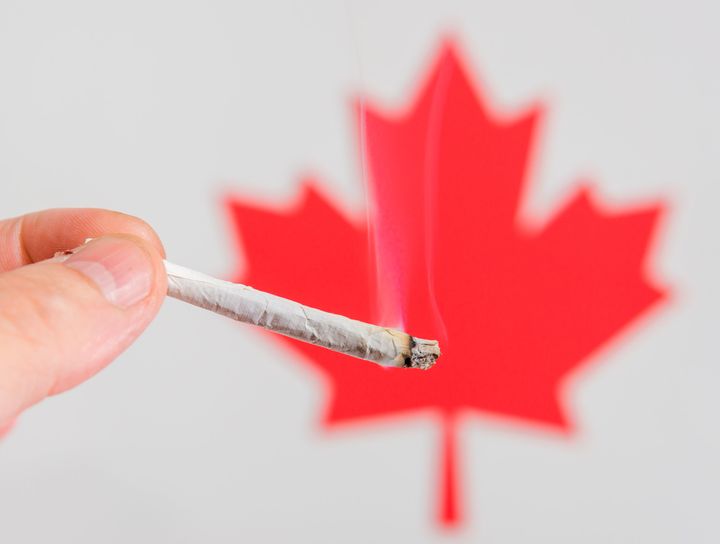 A hand holding a marijuana cigarette in front of a red maple leaf. The joint is lit and smoke rises from it. There is room for test above the joint.