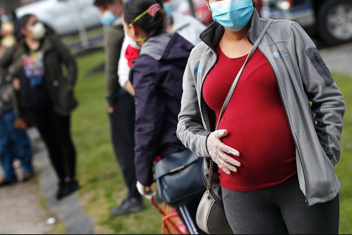 A pregnant woman wearing a face mask and gloves holds her belly as she waits in line for groceries at a food pantry in Waltha