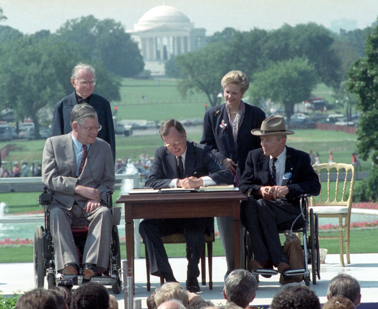 President George H.W. Bush signs the Americans With Disabilities Act on the South Lawn of the White House on July 26, 1990. With him are the Rev. Harold Wilke (rear left), Evan Kemp, chair of the Equal Opportunity Employment Commission (left), Sandra Parrino, chair of the National Council on Disability, and Justin Dart, chair of The President's Council on Disabilities. Jefferson Memorial is in the background.
