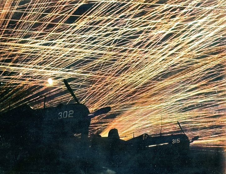 1945年4月28日、読谷飛行場にて。空襲する日本軍機に向けた対空砲火。アメリカ海兵隊の「Hell's Belles」飛行隊のF4U「コルセア」戦闘機がシルエットとして浮かび上がる。