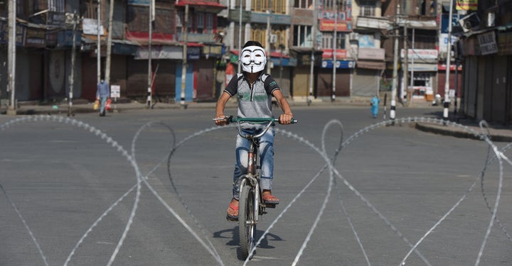 A boy wearing a Guy Fawkes mask bicycles near barbed wire laid out during curfew at the Sarie Bala area on August 4, 2020 in Srinagar, India.