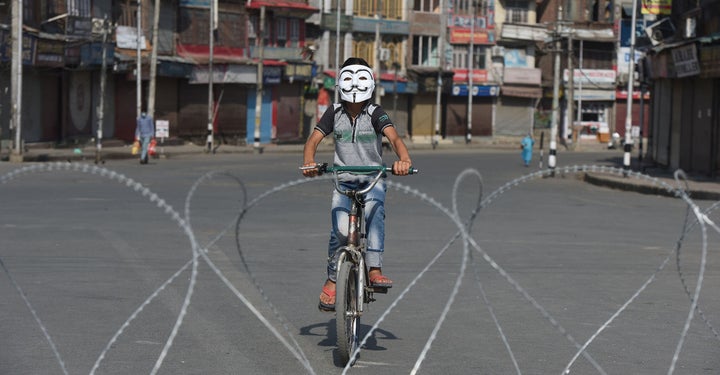 A boy wearing a Guy Fawkes mask bicycles near barbed wire laid out during curfew at the Sarie Bala area on August 4, 2020 in Srinagar, India. 