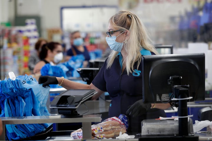 Cashier Mercedes Peña wears a protective mask as she works behind a plastic shield at the Presidente Supermarket this past April in Hialeah, Fla. All employees are required to wear masks which are provided by the company. (AP Photo/Lynne Sladky)