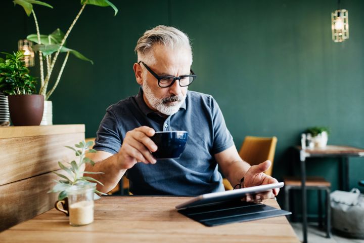 Man sitting in a cafe, reading on a digital tablet and drinking a cup of coffee.