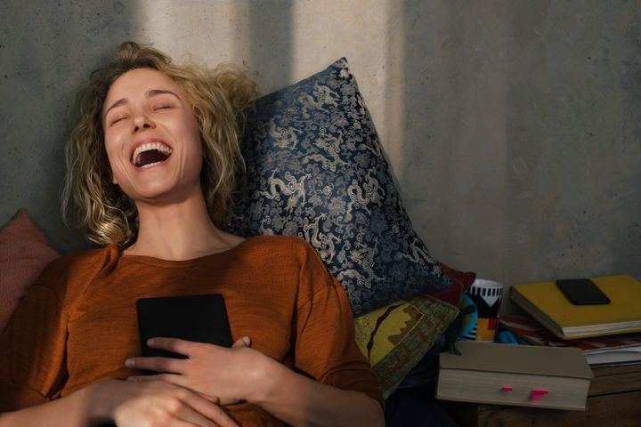 Portrait of laughing young woman lying on bed with E-book reader