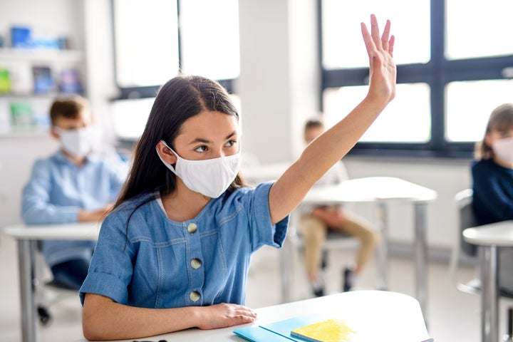 Girl with face mask back at school after covid-19 quarantine and lockdown, raising hand.