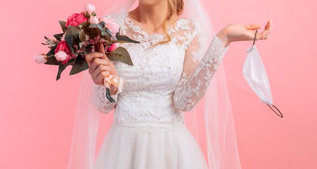 Bride in a wedding dress with a medical protective mask in her hands, on a pink background