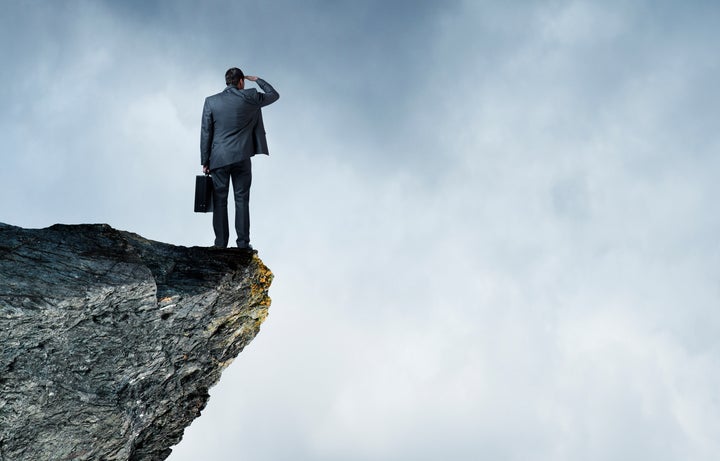 A businessman standing on the edge of a cliff and holding his briefcase, peers out towards a fog shrouded sea. Ample copy space is created by the clouds.