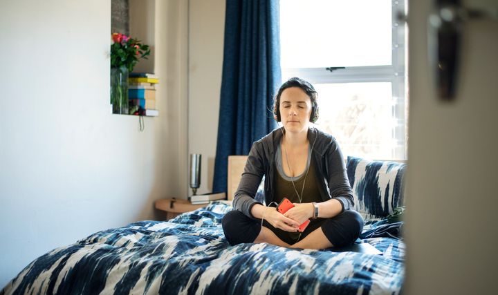 Young woman sitting on her bed with her eyes closed and listening to an audio meditation on headphones