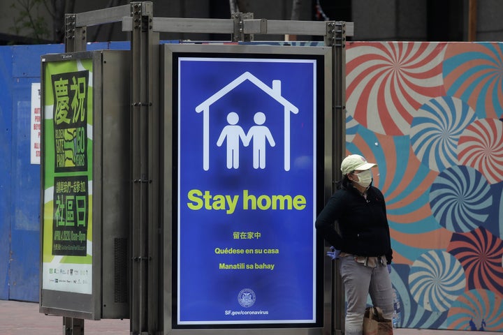 A woman wearing a mask walks past a sign advising people to stay home for concerns over the COVID-19 pandemic at a MUNI bus stop in San Francisco, Saturday, April 18, 2020. (AP Photo/Jeff Chiu)