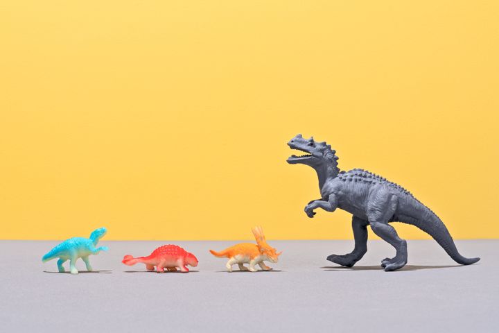 Small Toy Dinosaurs Facing A Large Toy Dinosaur on Yellow Colored Background.