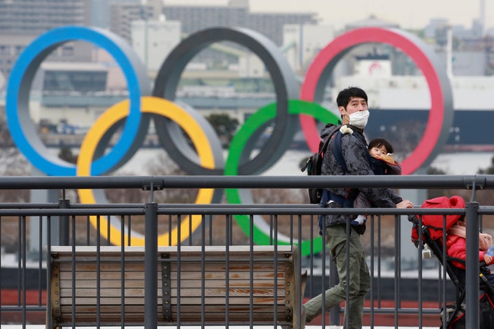 Une personne portant un masque marche près de la mer alors que des anneaux olympiques sont vus en arrière-plan à Tokyo, le mardi 26 janvier 2021.