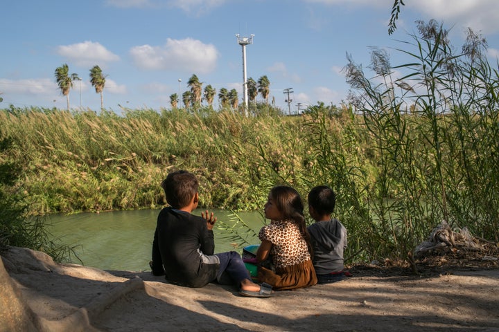 Young Central American asylum-seekers sit on the bank of the Rio Grande, as cameras from an American surveillance tower look