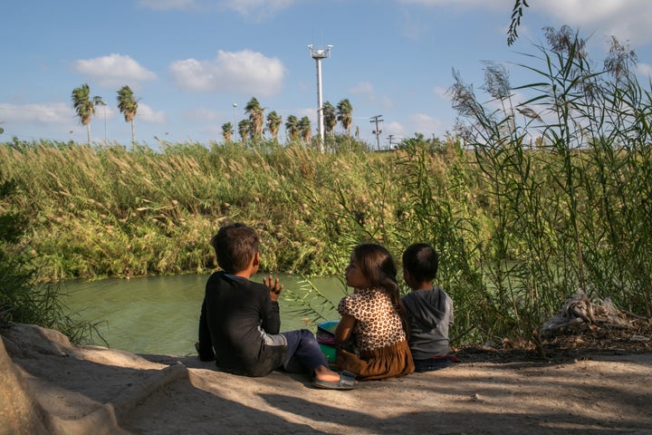 Young Central American asylum-seekers sit on the bank of the Rio Grande, as cameras from an American surveillance tower look on from across the border.