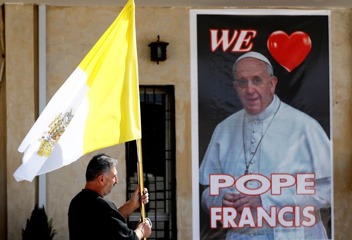 A priest holds a Vatican flag as he walks by a poster of Pope Francis during preparations for the Pope's visit in Mar Youssif