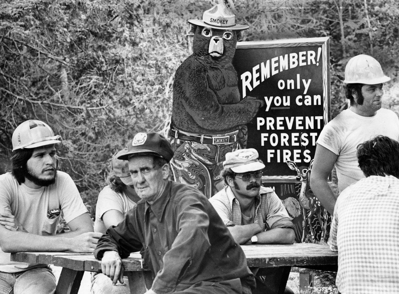 Smokey the Bear, the symbol of the nation's fire prevention efforts, stands watch at Togue Pond headquarters behind new crew awaiting transportation to fire lines of a forest fire in Millnocket, Maine, in 1977.