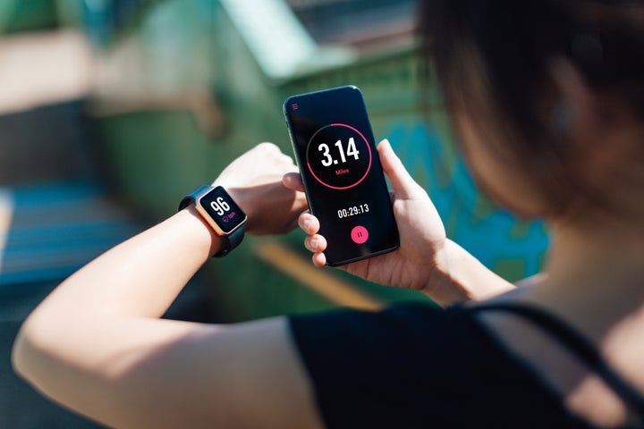 Over the shoulder view of a young woman holding smart phone and wearing smart watch, using fitness app to check her training progress.