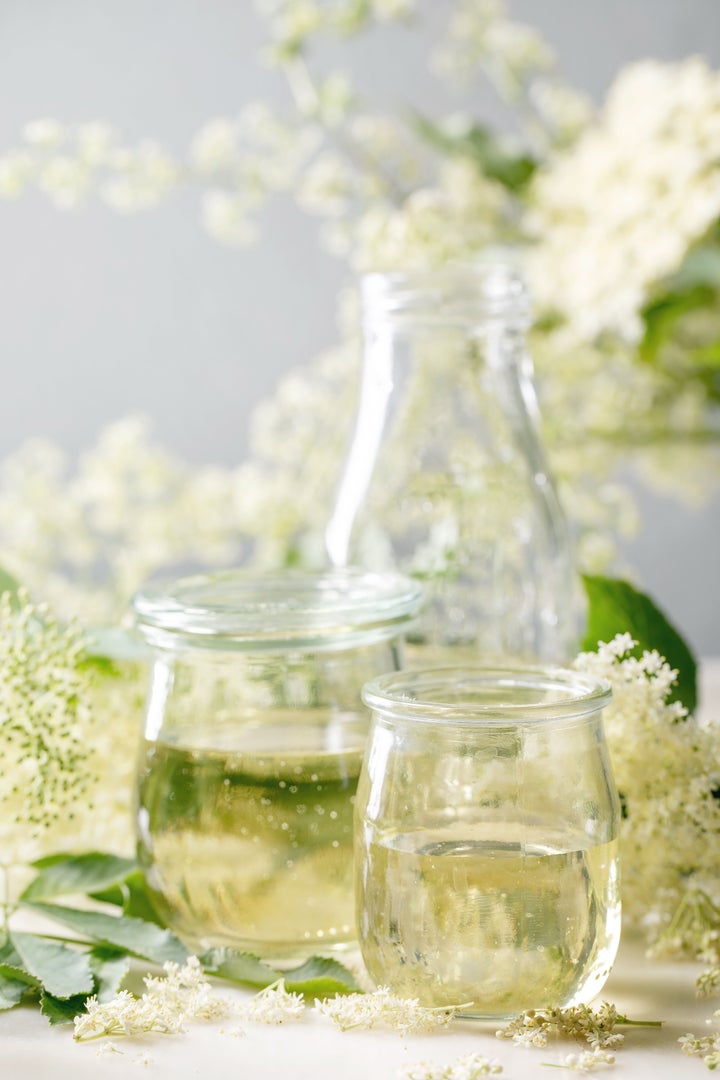 Elderberry flowers and leaves, glass jars of sweet homemade elderberry syrup on white marble table.