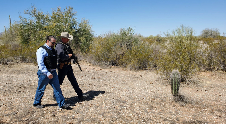 Arizona Attorney General Mark Brnovich, wearing a bulletproof vest, walks alongside an armed officer during a trip to the U.S.-Mexico border.