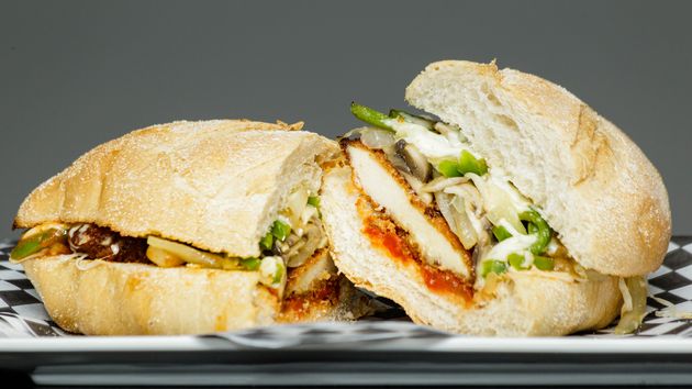 Chicken parm sandwich on a plate against a gray background. Some of the ingredients include chicken, onions, green peppers, mozzarella