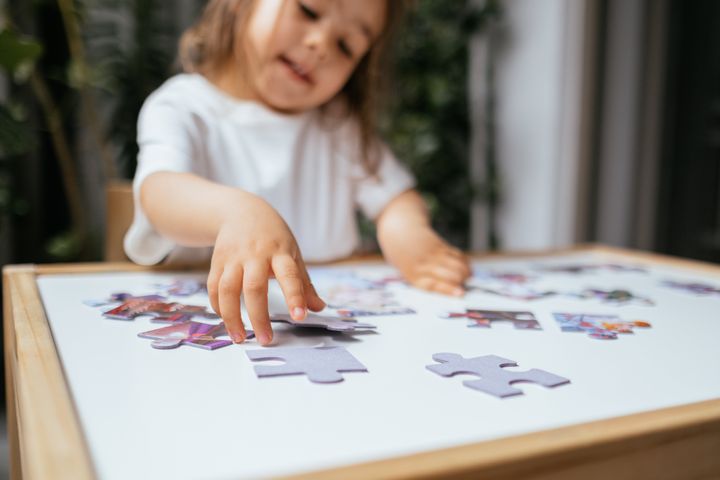 Little girl is trying to connect couple jigsaw puzzle piece. Side view of concentrated little girl playing with puzzles at home