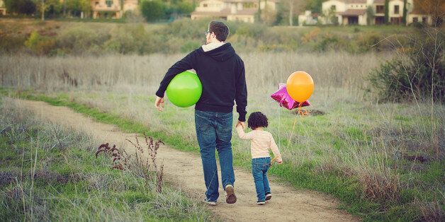 Father holding his little girl's hand as they walk together along a country trail. There are suburban homes in the background. She has colorful balloons.