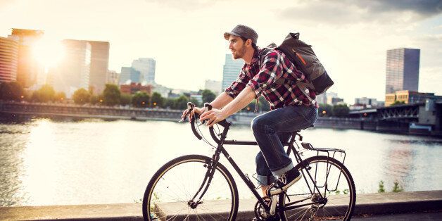 A young man commuting in an urban city environment on his street bicycle, a waterproof bike bag on his back. This is the Eastbank Esplanade in Portland, Oregon, that follows along the Willamette river. Downtown is visible across the water, the sun shining brightly between the buildings. Horizontal with copy space. In-camera lens flare.