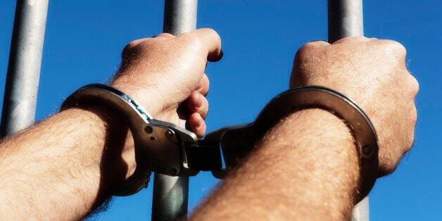 A handcuffed man's fists clutch the steel bars of a prison cell. Outside is freedom, represented by a clear blue sky.